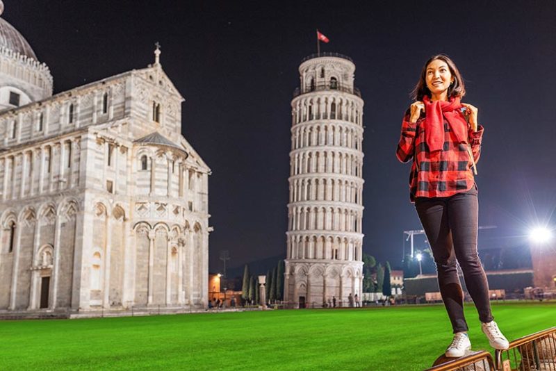Portrait of happy young woman tourist traveler in front of leaning tower of Pisa, Italy at evening time